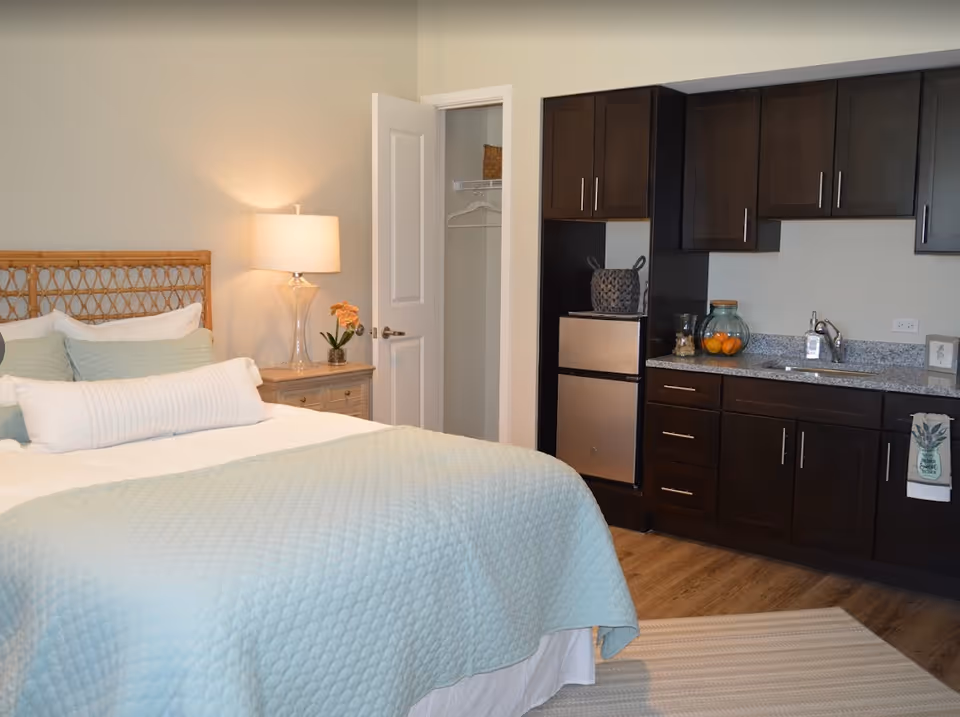 A cozy bedroom featuring a bed with light blue quilted bedding and multiple pillows. To the left of the bed is a wooden nightstand with a clear glass lamp and a small plant with orange flowers. Behind the bed is a partially open door revealing a small closet with a hanging rod and a basket on the shelf. On the right side of the room, there is a kitchenette with dark wood cabinets, a small refrigerator, a granite countertop with a sink, and decorative items including a glass jar with oranges and a towel hanging on the cabinet handle.