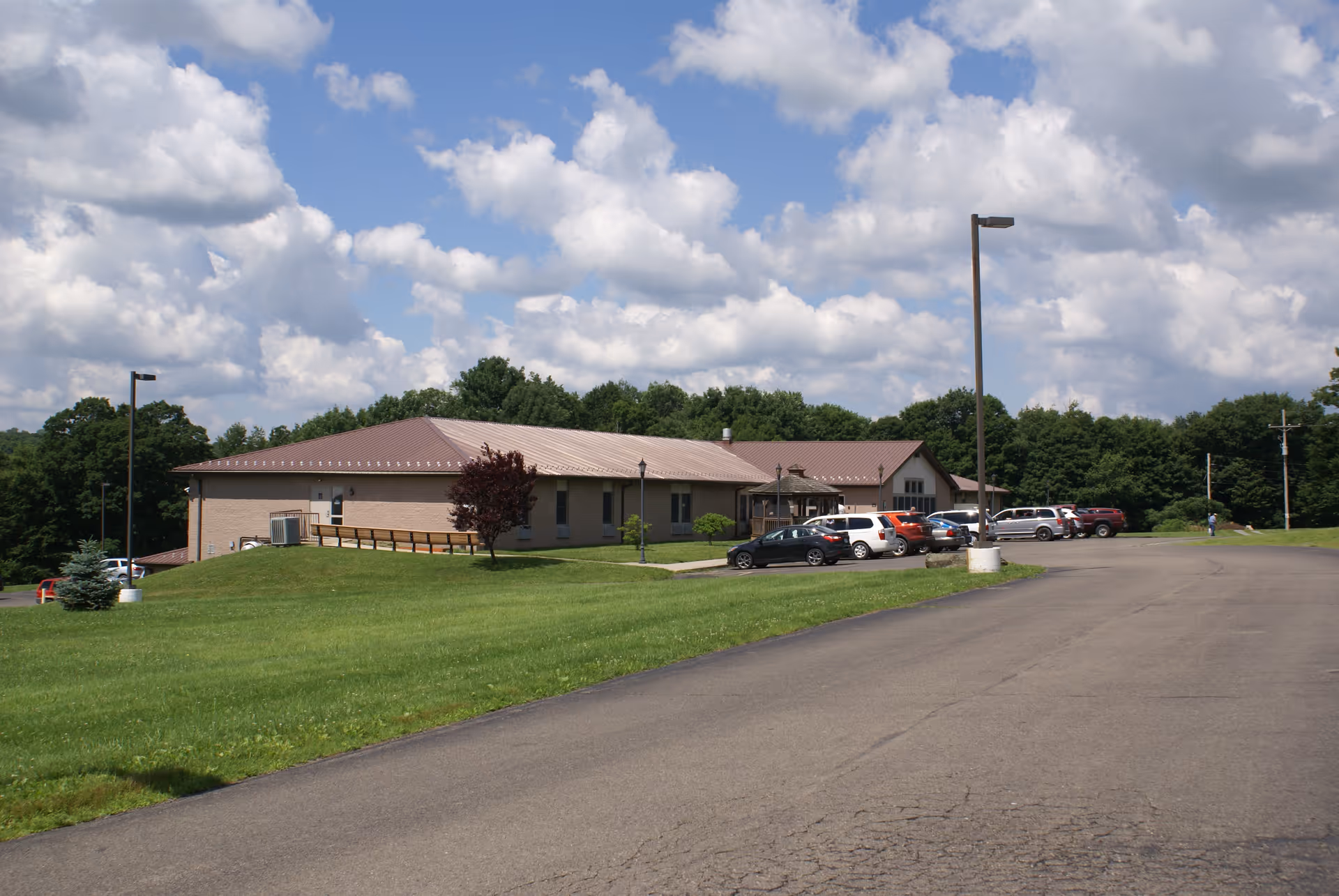 Single-story senior living building with a parking lot, lamp posts, and a large grassy lawn under a partly cloudy sky.