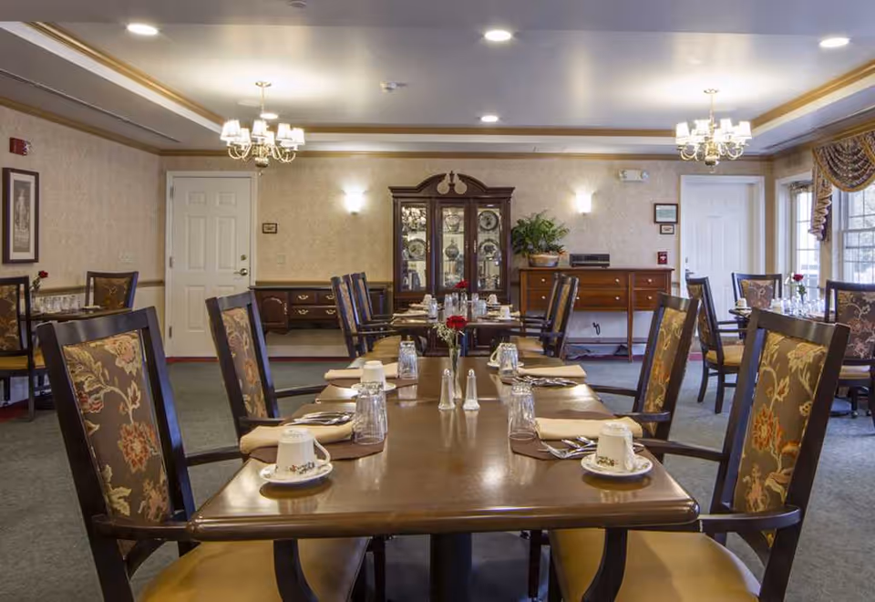 Formal dining room with long wooden tables set with cups, utensils, and floral-upholstered chairs.