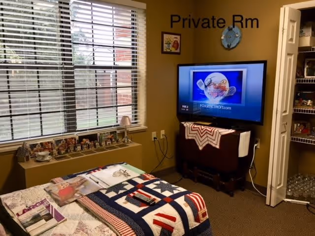A cozy private room with a bed covered in a colorful quilt, a window with blinds letting in natural light, a TV mounted on a stand displaying a FOX 2 Detroit screen, and an open closet with shelves stocked with various items including bottled water.