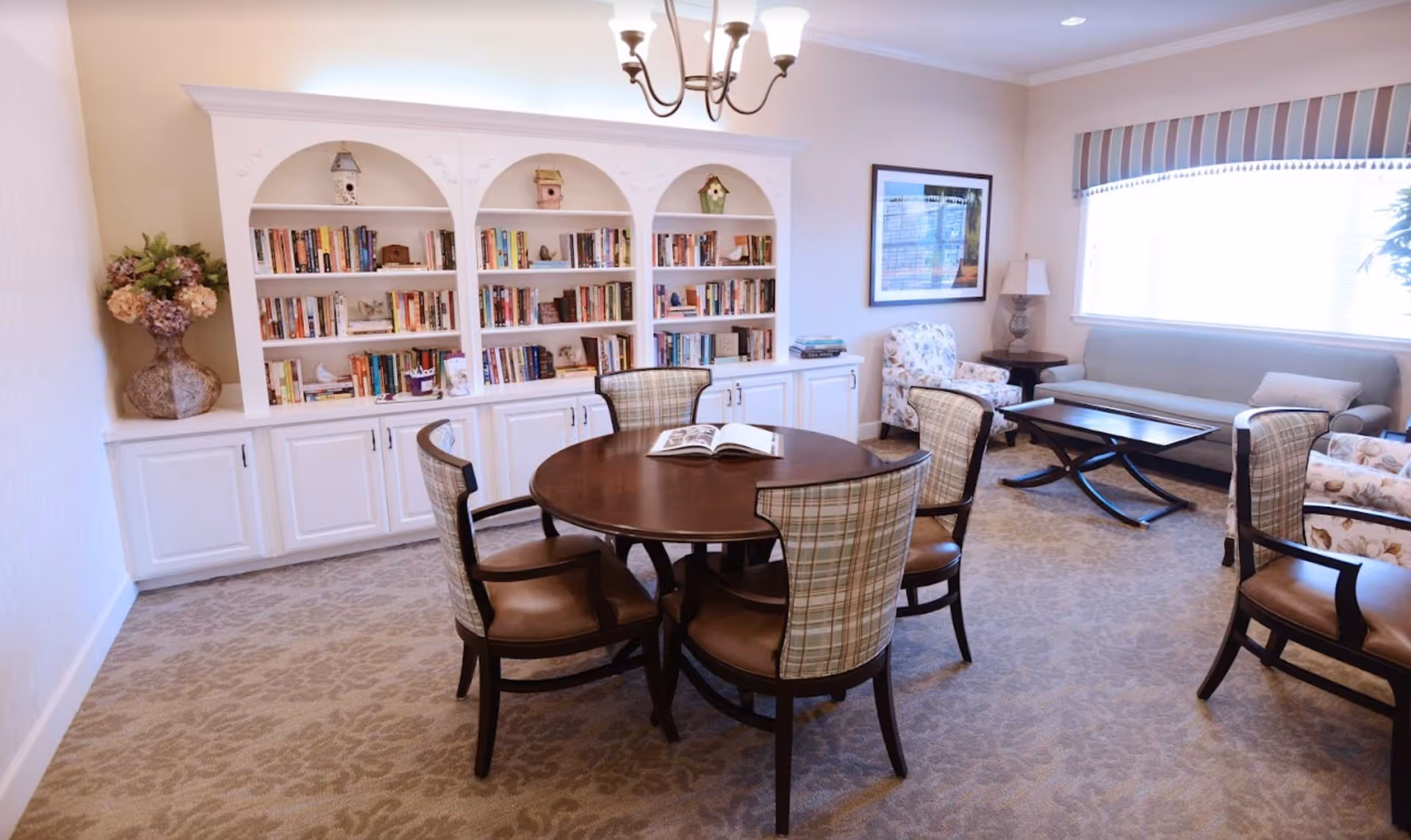 A cozy living room area with a round wooden table surrounded by five upholstered chairs with plaid backs. Behind the table is a white built-in bookshelf filled with books and decorative birdhouses. To the right, there is a seating area with a sofa, two armchairs with floral patterns, a coffee table, a side table with a lamp, and a large window with a striped valance allowing natural light to fill the room. A chandelier hangs from the ceiling.