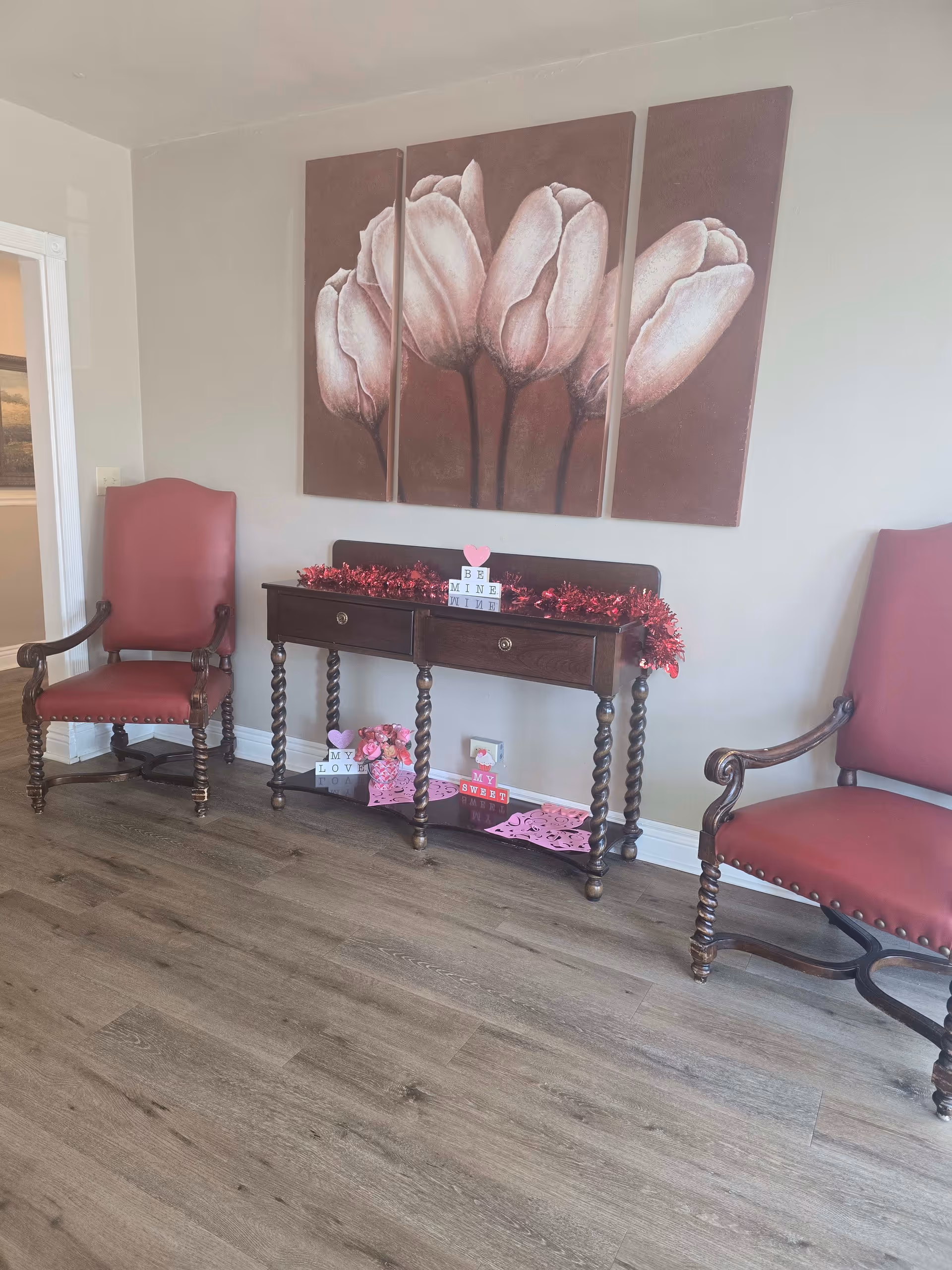 A room with two red upholstered wooden armchairs on either side of a dark wooden console table with twisted legs. The table is decorated with red garland and Valentine's Day themed blocks and decorations. Above the table is a four-panel painting of large white flowers on a reddish-brown background. The floor is light wood, and the walls are painted light gray.