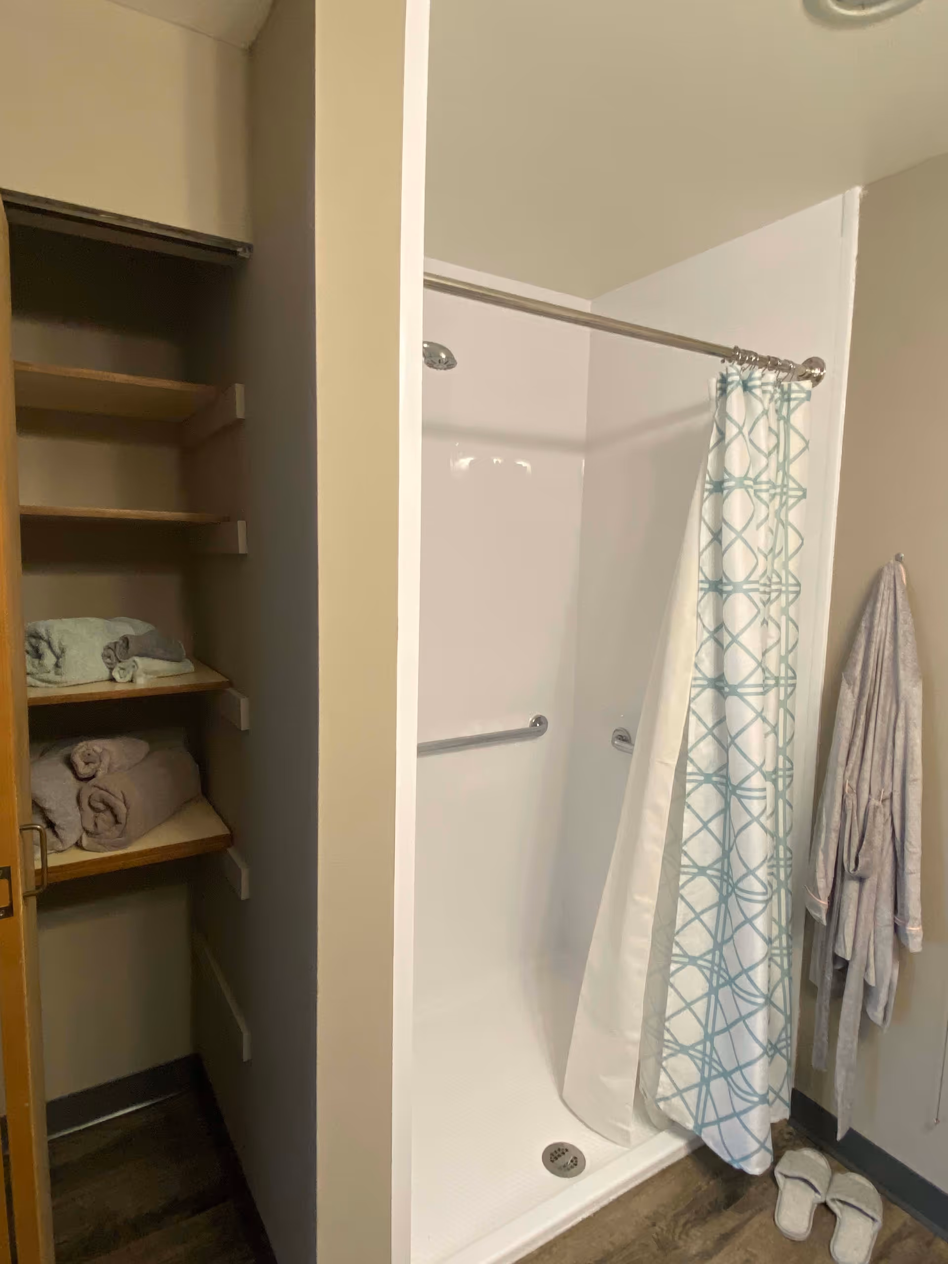 A bathroom shower area with a white shower stall, a silver shower curtain rod holding a white curtain with a blue geometric pattern. To the left, there is an open shelving unit with folded towels. On the right wall, a bathrobe hangs next to a pair of white slippers on the floor.