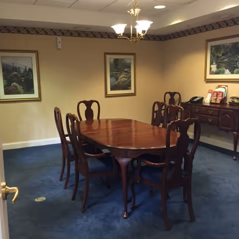 A dining room with a polished wooden table surrounded by six matching wooden chairs with blue cushions. The room has beige walls adorned with three framed landscape paintings and a decorative border near the ceiling. A wooden sideboard with brochures and decorative items is against one wall, and a chandelier with five lights hangs above the table. The floor is covered with blue carpet.