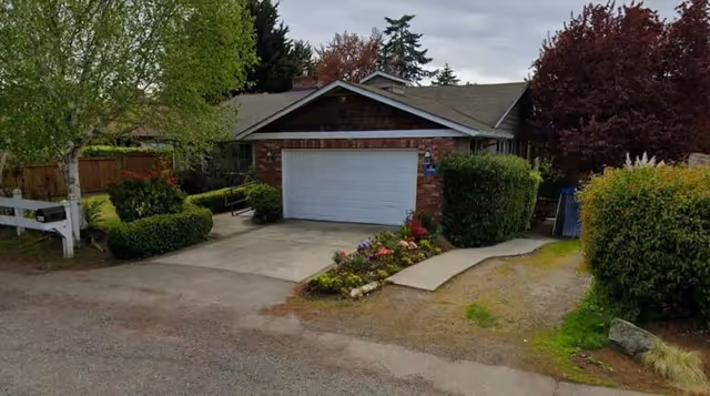 Front exterior of a single-story brick house with a two-car garage, driveway, and landscaped yard with trees and shrubs.