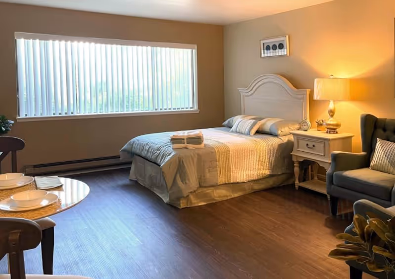 Sunlit studio bedroom with a made bed, nightstand and lamp, armchair, and a small dining table by a large window with vertical blinds.