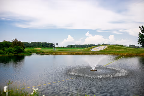 A peaceful outdoor scene featuring a pond with a water fountain in the center, surrounded by green grass and trees under a partly cloudy sky. In the background, there is a grassy hill with a large circular clock embedded in it.