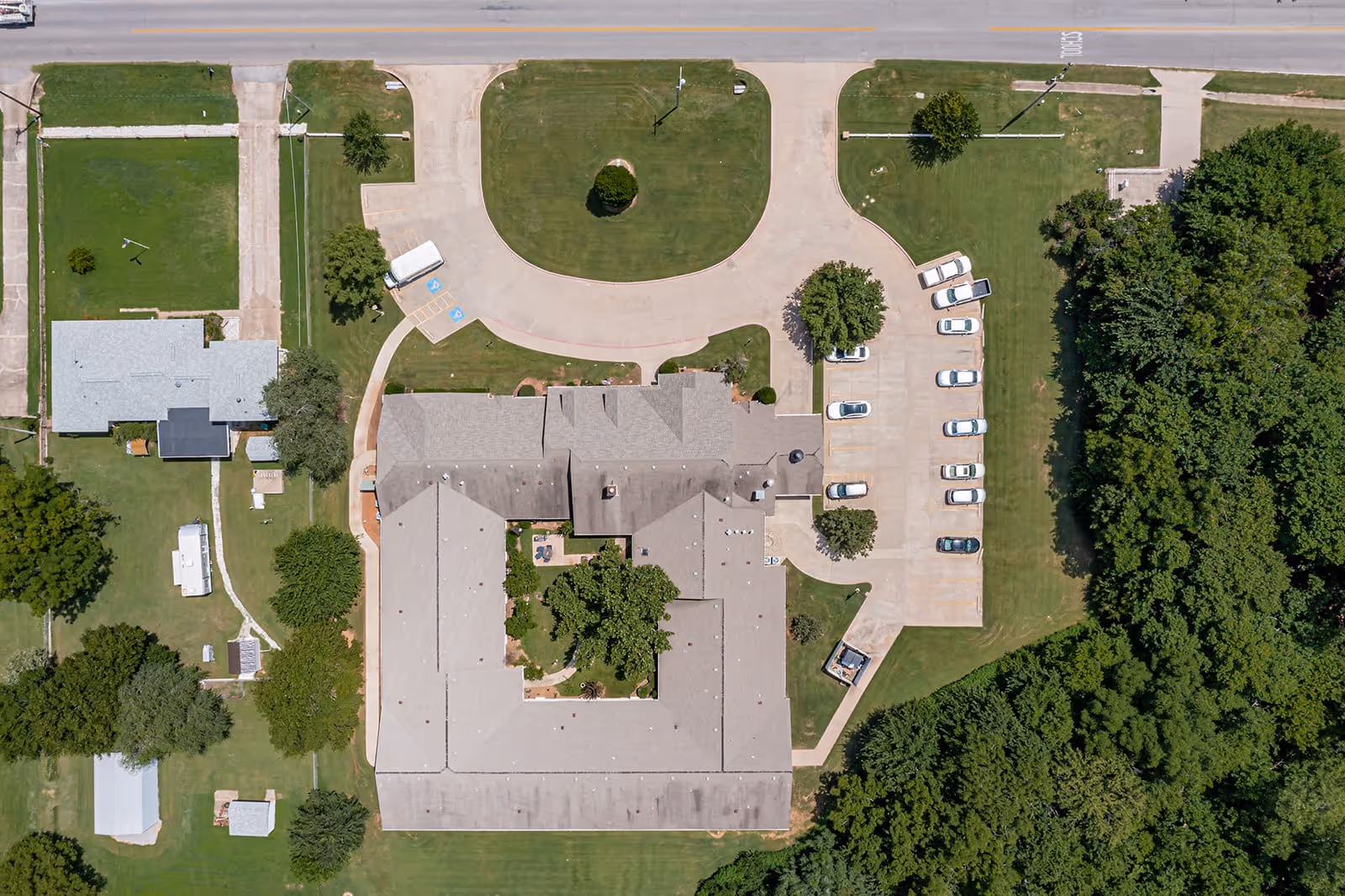Aerial view of Ashbrook Village facility showing a large building with a U-shaped layout surrounded by green lawns, trees, parking spaces with several cars, and adjacent smaller buildings. A road runs along the top edge of the image.