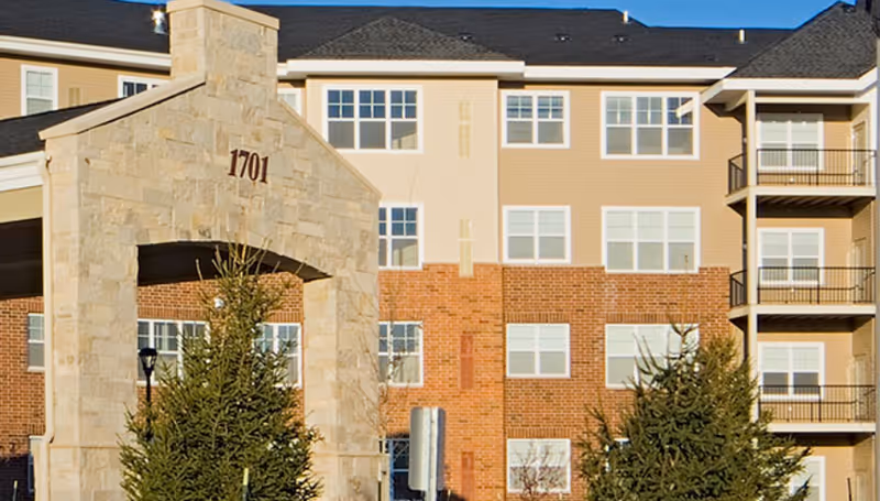 Stone entry arch marked 1701 in front of a multi-story building with brick and siding facade, balconies, and evergreen trees.