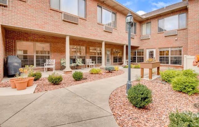 Outdoor courtyard area of a senior living facility with a brick two-story building surrounding it. The courtyard features a concrete walkway, landscaping with small bushes and plants, a lamp post, a foosball table, and several white chairs arranged near the building windows.