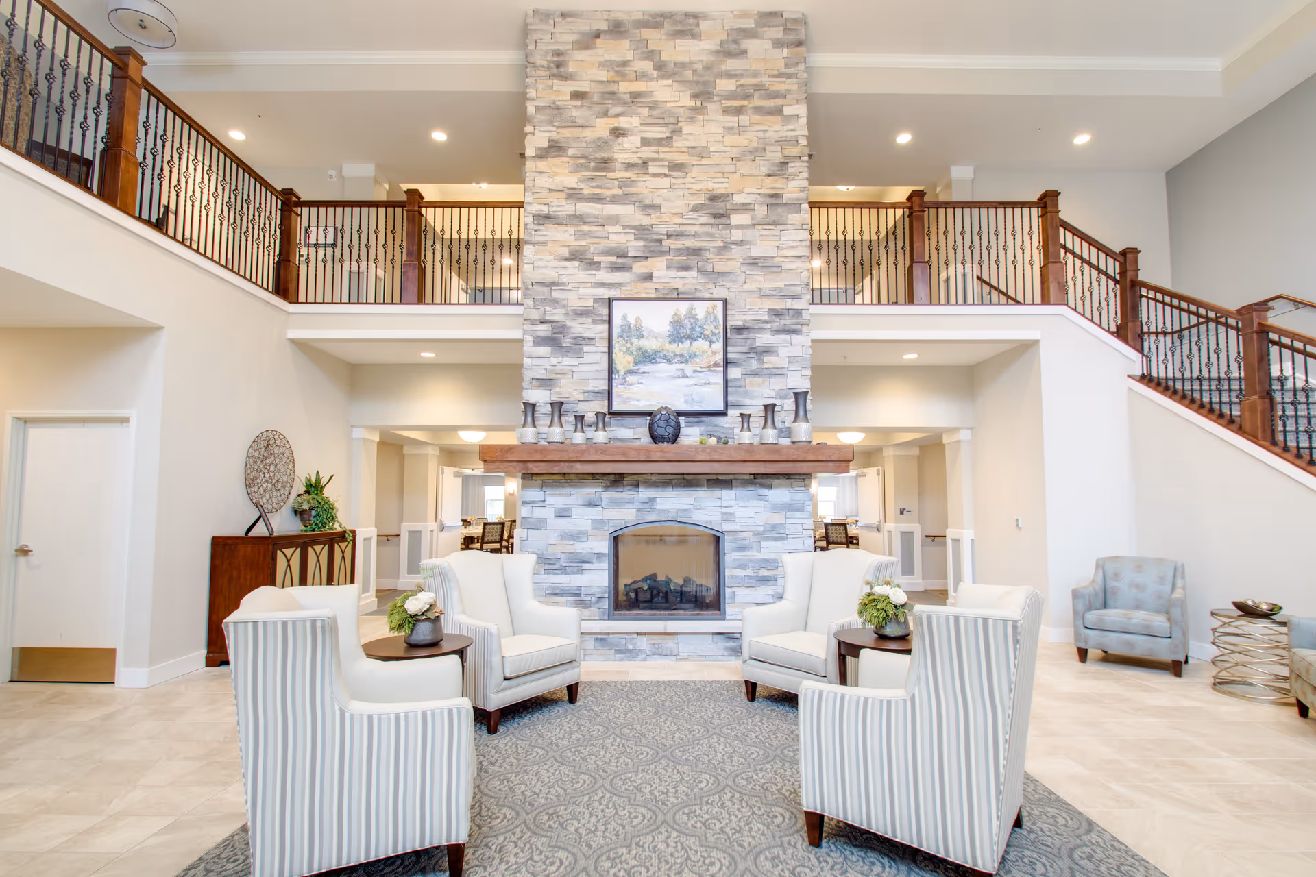 A spacious and bright senior living facility common area featuring a large stone fireplace with a wooden mantel. Four upholstered armchairs arranged around two small round tables with flower arrangements sit on a patterned rug in front of the fireplace. The room has high ceilings with a second-floor balcony railing overlooking the area. A staircase with wooden handrails is visible on the right side, and additional seating and decorative elements are placed around the room.