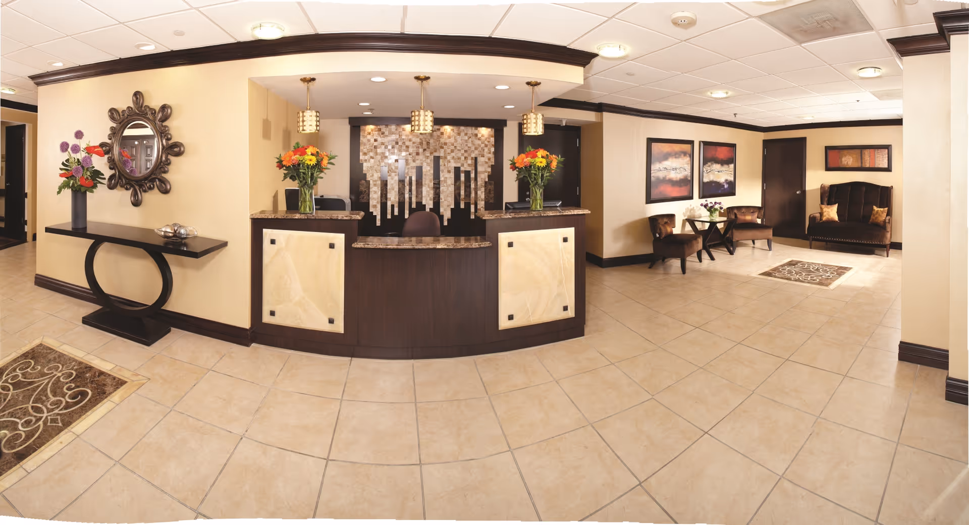 Lobby reception area with a dark wood front desk, tiled floor, floral arrangements, and seating with artwork on the walls.
