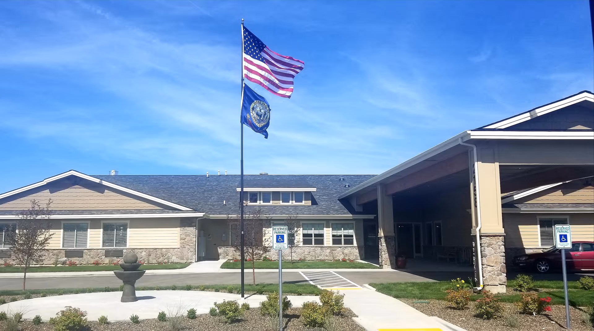 Exterior view of a single-story senior care facility building with a covered entrance, stone and beige siding, two flags on a flagpole, and reserved parking signs for handicapped parking spaces in front. The sky is clear and blue.