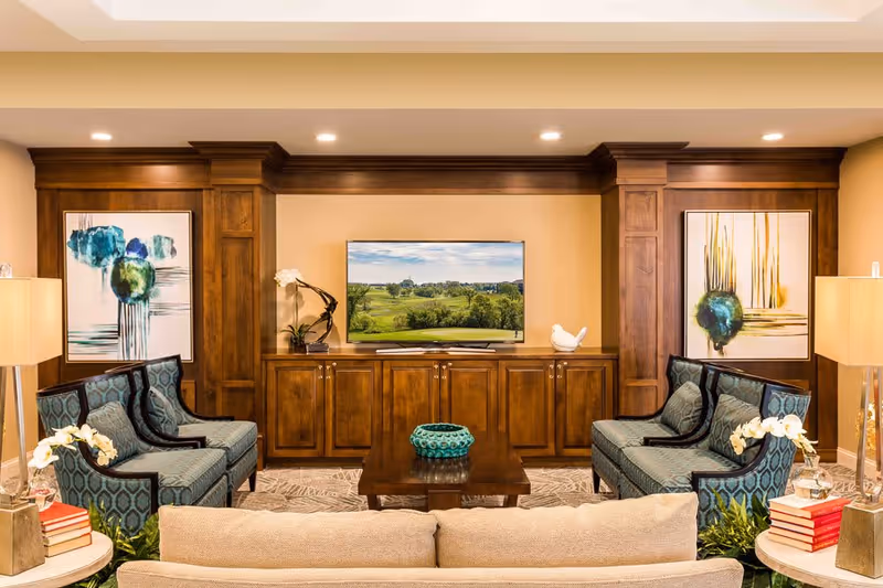 A cozy living room with two patterned blue armchairs facing a wooden coffee table and a flat-screen TV mounted on a wooden cabinet. The room features warm wood paneling, two large abstract paintings on the walls, table lamps, and decorative plants and books on side tables.