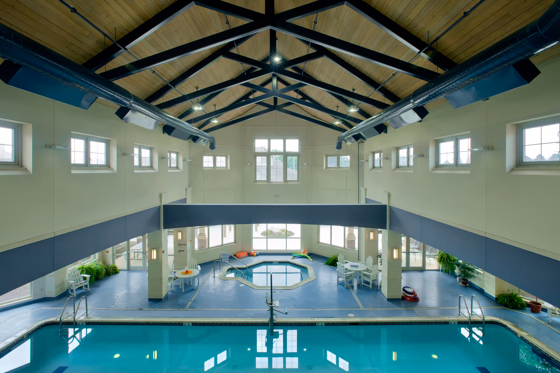 Bright indoor swimming pool and hot tub in a spacious recreation room with a vaulted wood-beamed ceiling and windows.