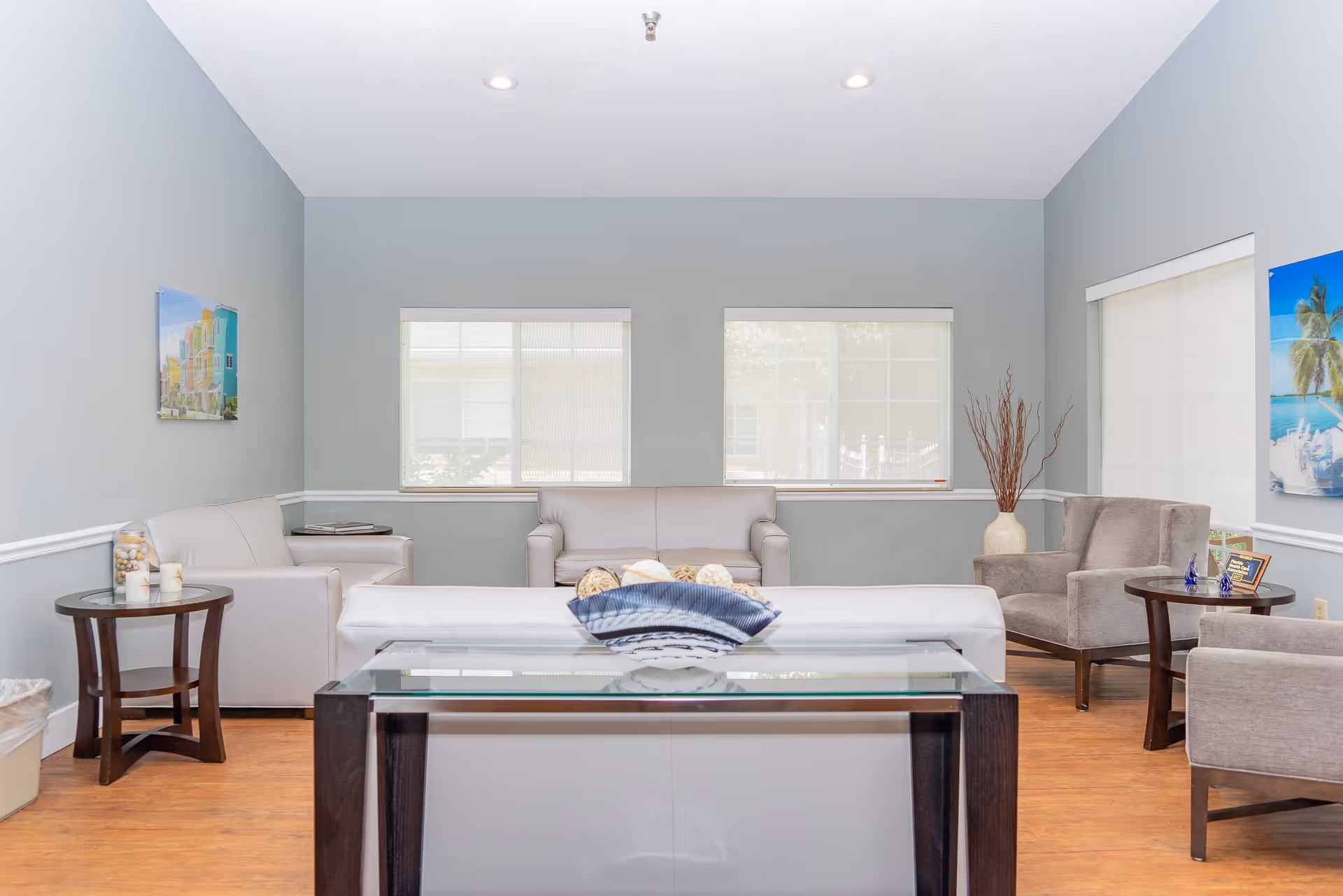 A bright and clean living room area with light gray walls and wooden flooring. The room features a white leather sofa, two gray armchairs, a glass coffee table with decorative items, and side tables with small decorations. There are three windows with white blinds letting in natural light, and two colorful framed pictures on the walls.