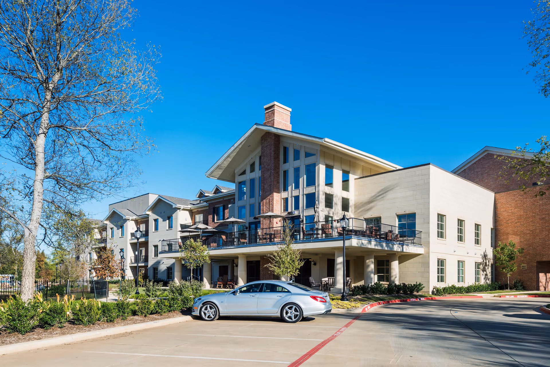 Exterior view of a modern senior living facility building with large windows, a balcony with outdoor seating, and a silver car parked in front. Trees and landscaping surround the building under a clear blue sky.