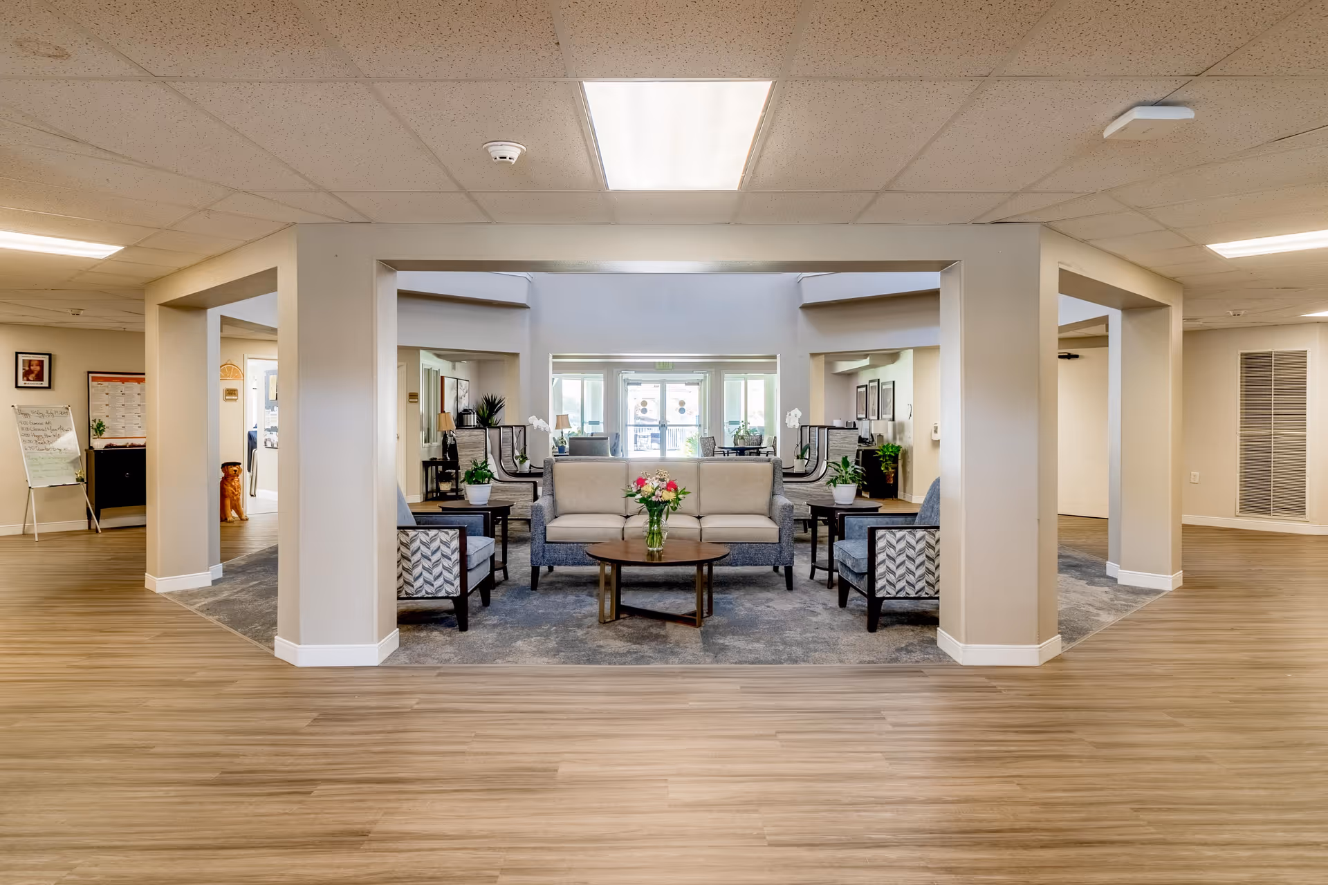 A spacious and well-lit common area in a retirement community featuring a seating arrangement with a beige sofa, patterned armchairs, and a round wooden coffee table with a vase of flowers. The area is framed by large square columns and has wood flooring with a carpeted section under the seating. In the background, there are large windows and glass doors letting in natural light, along with plants and framed pictures on the walls.