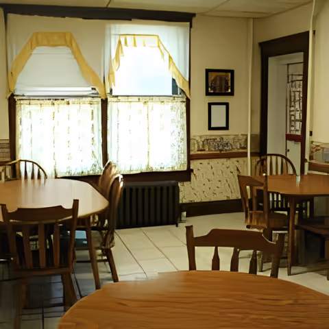 A dining room with several wooden tables and chairs. The room has two windows with white and yellow curtains, a radiator below the windows, and framed pictures on the wall. The floor is tiled and there is a door with a glass panel on the right side.