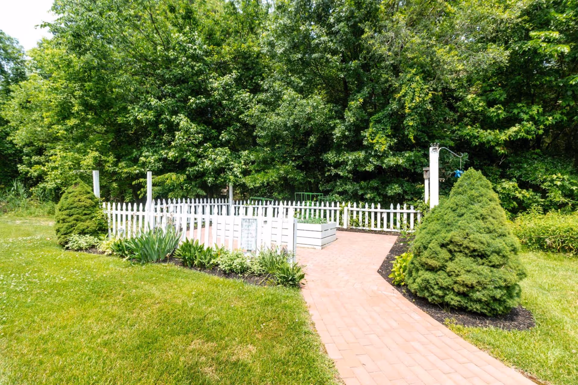 Brick pathway leading to a white picket-fenced garden area surrounded by shrubs and trees.