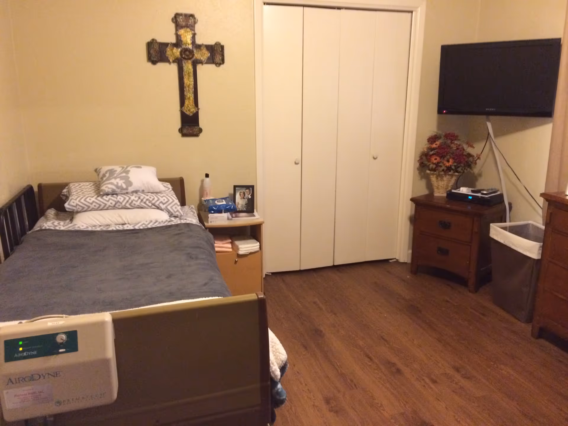 A small bedroom with a single bed covered with pillows and a gray blanket. Above the bed on the wall is a decorative cross. Next to the bed is a small nightstand holding personal items including a photo frame and a box of wipes. Across from the bed is a wooden dresser with a basket of artificial flowers and a flat-screen TV mounted on the wall above it. The room has wooden flooring and a closed white closet door.