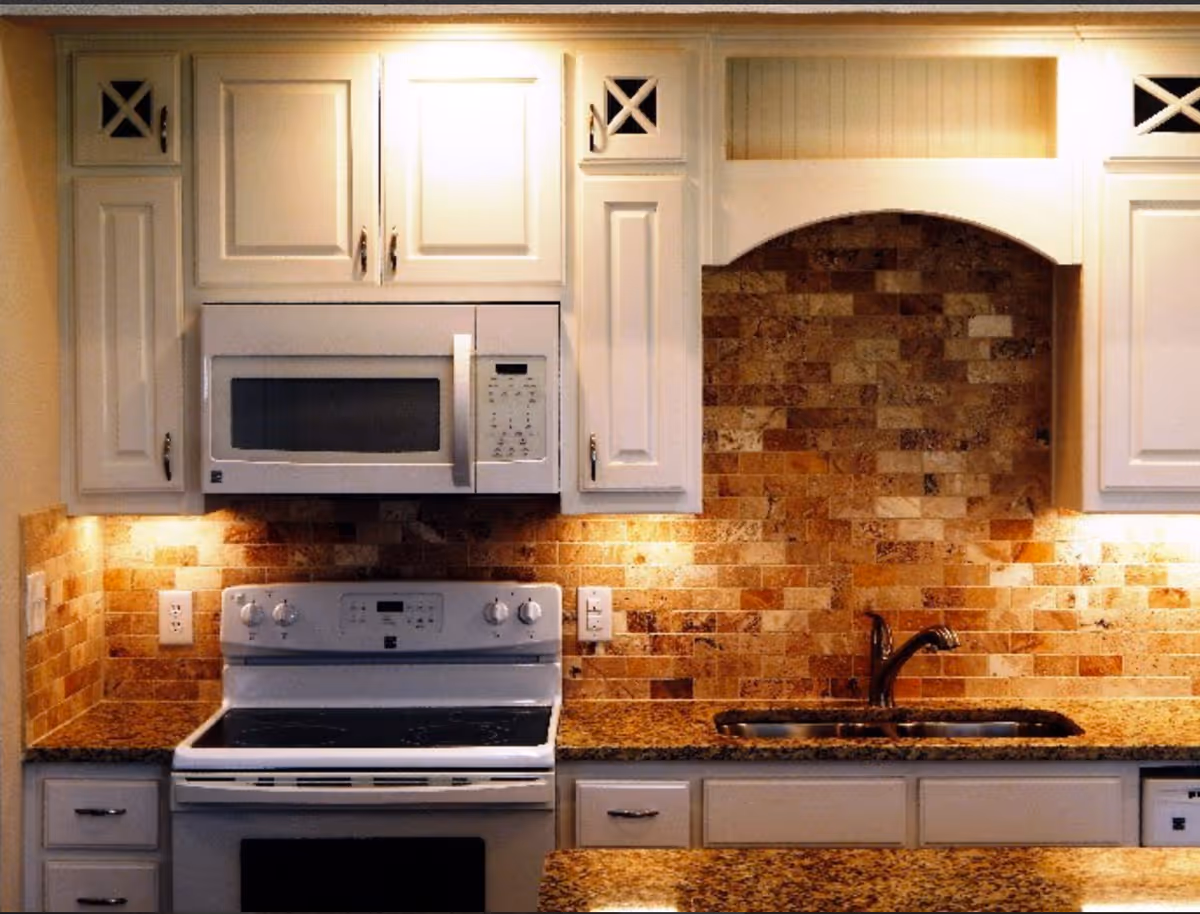 A kitchen interior featuring white cabinets, a white microwave above a white electric stove, a granite countertop, a stainless steel sink with a faucet, and a backsplash made of brown and beige stone tiles.