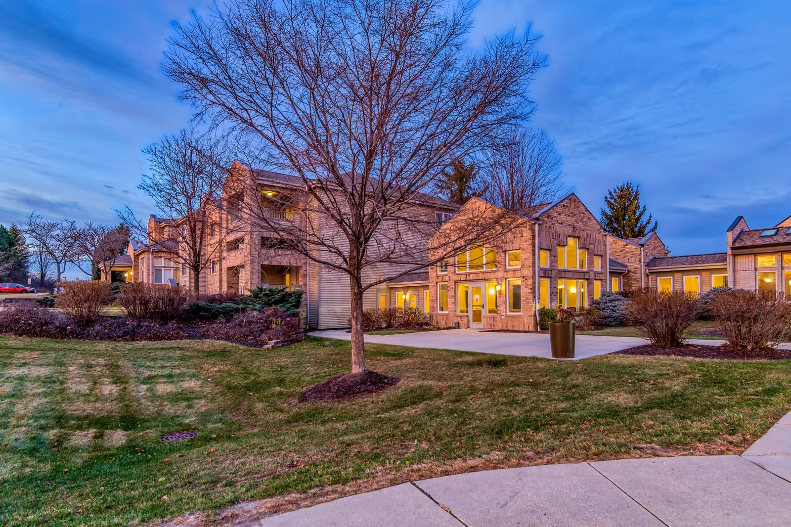 Exterior view of Harbour House senior living facility at dusk, showing a multi-story building with large windows illuminated from inside, surrounded by leafless trees and landscaped bushes on a grassy lawn.