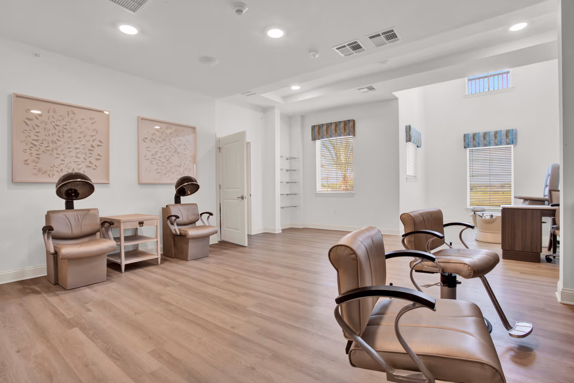 A bright and spacious salon area with four brown leather styling chairs, two hair dryer chairs, a small wooden side table, and large windows with blinds and valances letting in natural light. The room has light wood flooring and white walls with two framed artworks.