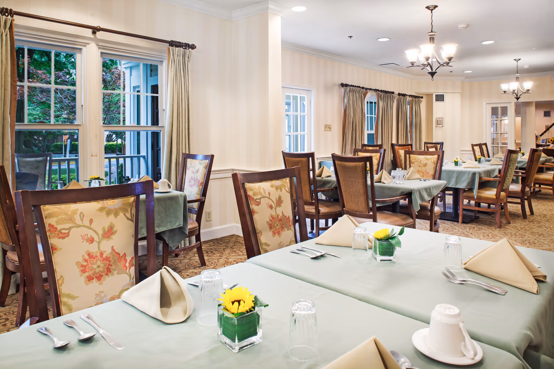 Bright dining room with multiple tables set with folded napkins, place settings, small flower centerpieces, and wooden chairs near windows.
