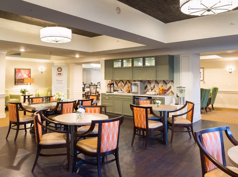 Communal dining area with round tables, orange-patterned chairs, and a small serving counter and kitchenette in the background.