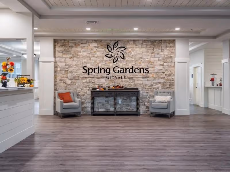 Interior view of a senior living facility lobby with a stone accent wall featuring the Spring Gardens Midvale logo. Two gray armchairs with cushions flank a dark wooden console table with decorative items. The space has wood flooring, white walls, and a ceiling with recessed lighting.