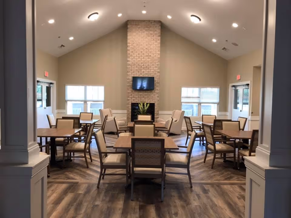 A spacious dining room in a senior living facility with multiple wooden tables and cushioned chairs arranged neatly. The room features a high vaulted ceiling, a brick fireplace with a mounted flat-screen TV above it, and large windows letting in natural light. The flooring is wood-patterned, and the walls are painted in a neutral tone.