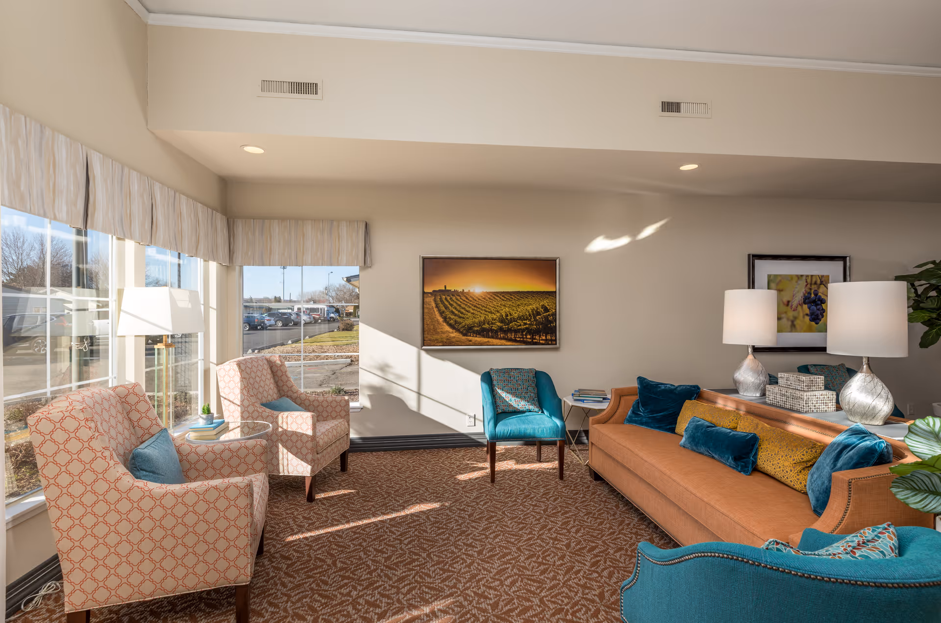 A bright and cozy living room area with large windows letting in natural light. The room features a brown sofa with blue and yellow cushions, two patterned armchairs with blue pillows, and a teal armchair. There are two table lamps on a console table behind the sofa, and framed artwork on the walls. Outside the windows, a parking lot and some trees are visible.