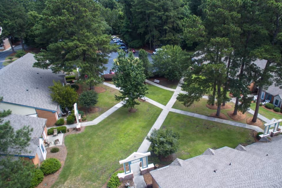 Aerial view of a senior living facility courtyard with green lawns, trees, and intersecting concrete walkways surrounded by single-story buildings with gray roofs.