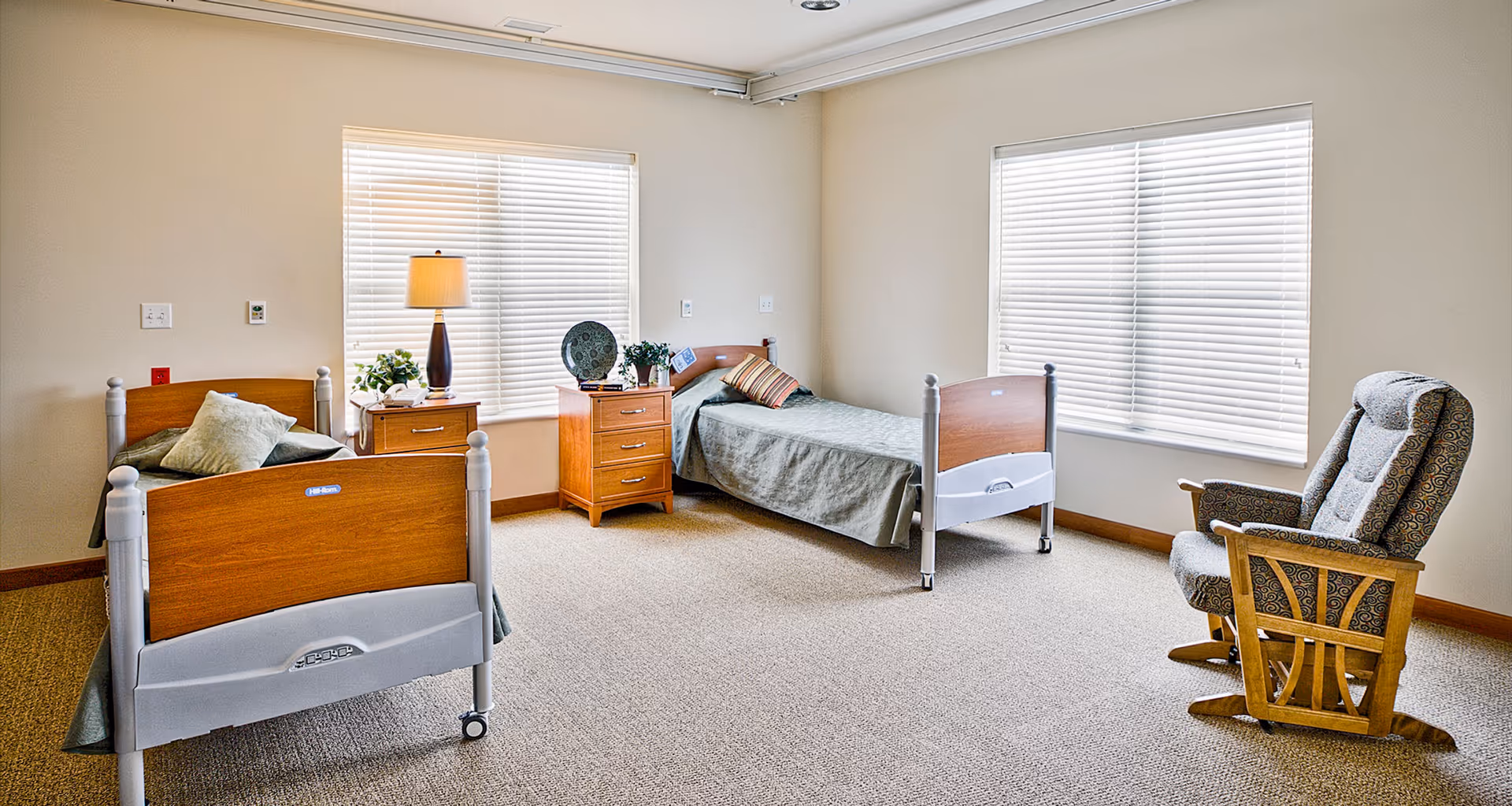 A bright shared senior bedroom with two single beds, wooden bedside tables, a lamp, and a patterned recliner by the windows.