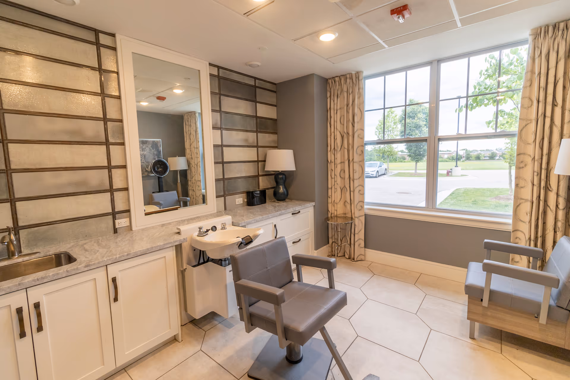 Interior of a salon area in a senior living facility with a salon chair facing a wash basin and a large mirror on the wall. The room has a tiled floor, a countertop with cabinets, a sink, a lamp, and large windows with curtains showing a view of trees and parked cars outside.