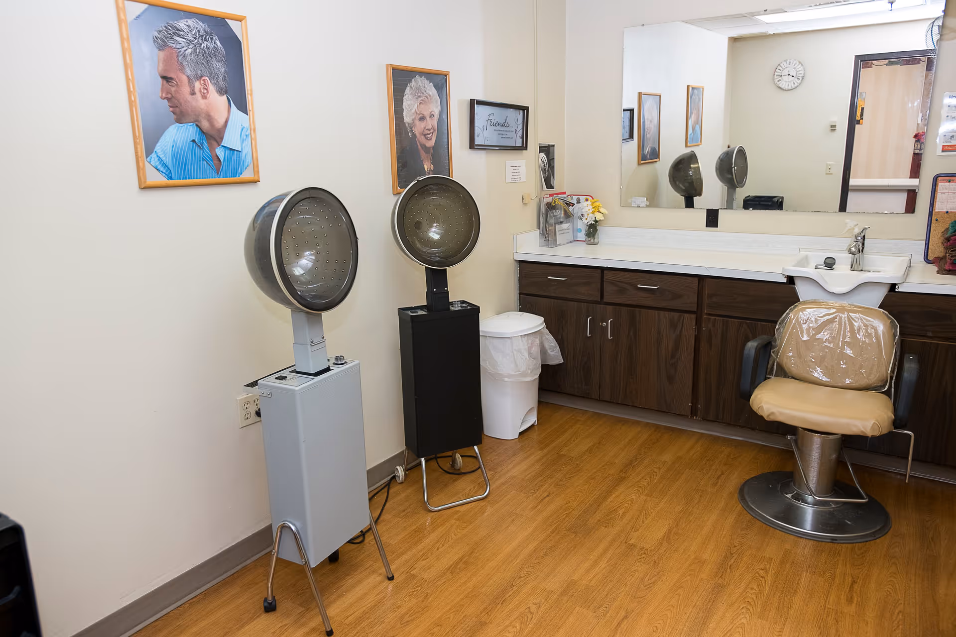 Interior of a hair salon area in a senior living facility with two vintage hair dryers, a salon chair covered in plastic, a large mirror above a counter with cabinets, and framed photos on the wall.