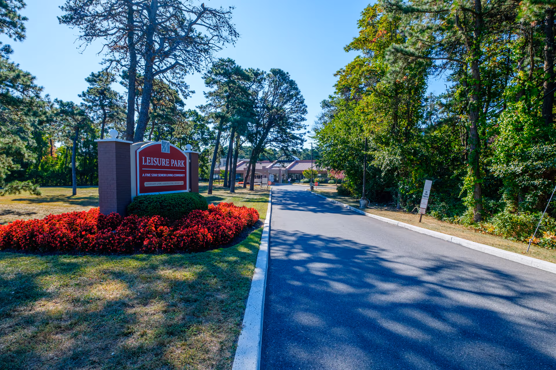 Entrance to Leisure Park senior living community with a red sign surrounded by red flowers on the left and a paved driveway leading to the building in the background, bordered by trees and greenery on both sides under a clear blue sky.