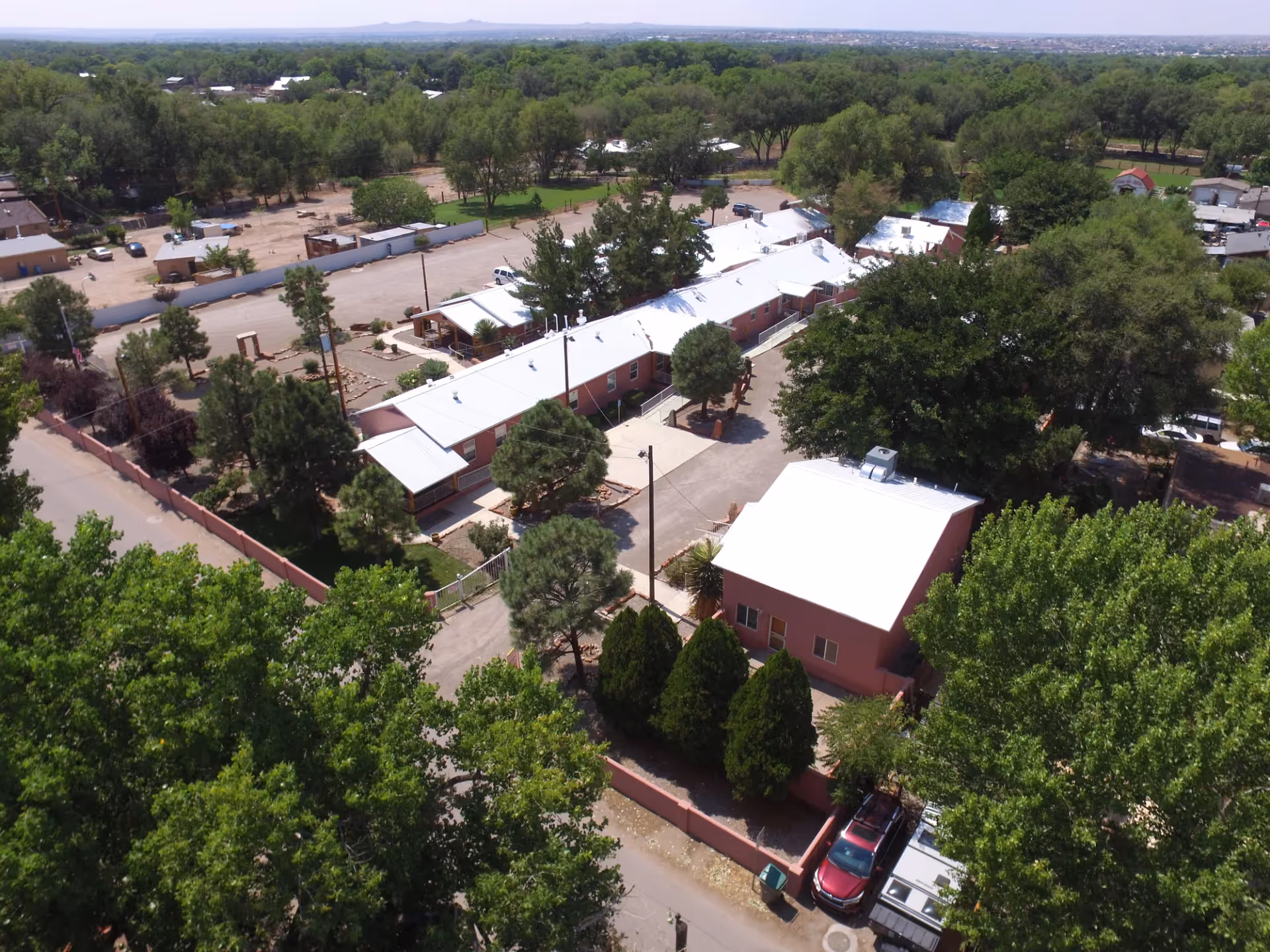 Aerial view of Casa de Rosa Assisted Living facility showing multiple single-story buildings with white roofs surrounded by trees and greenery. The area includes paved roads, parking spaces, and a mix of open and wooded spaces extending into the distance.