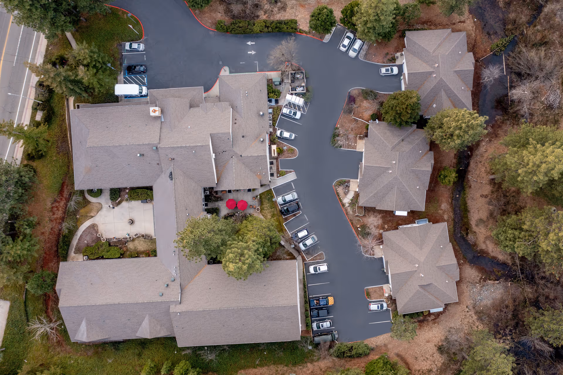 Aerial view of Cascades of Grass Valley senior living facility showing multiple buildings with gray roofs, parking spaces with cars, surrounding trees, and a small creek running alongside the property.