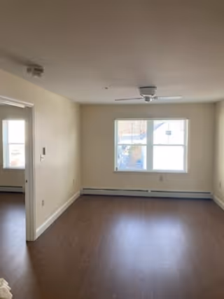 Empty room with light beige walls, a window with white trim letting in natural light, a ceiling fan with light fixture, and dark wood flooring. There is an open doorway leading to another room with a similar window and flooring.