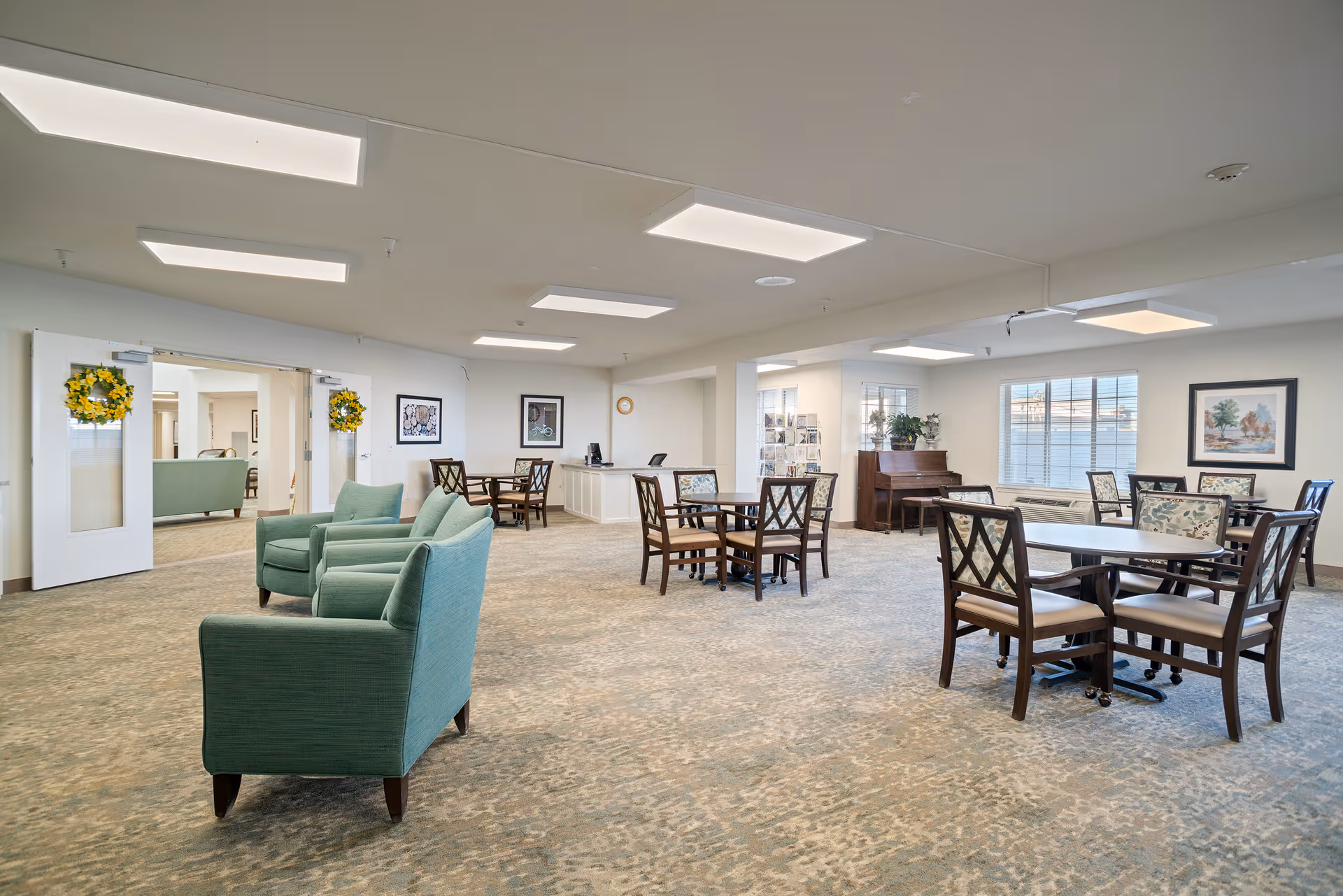 Bright common room with green armchairs, several dining tables and chairs, and a piano in a retirement facility.