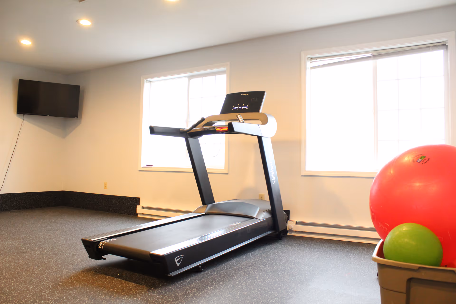 A small fitness room with a treadmill positioned in front of two large windows. A wall-mounted TV is visible on the left side, and a container holding a large red exercise ball and a smaller green ball is on the right side.