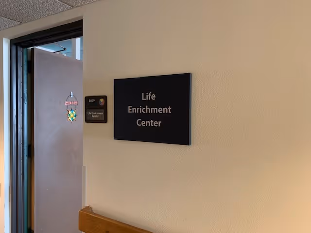 A beige wall with a black sign that reads 'Life Enrichment Center' next to an open door. The door has a colorful hanging decoration with the word 'Welcome'.