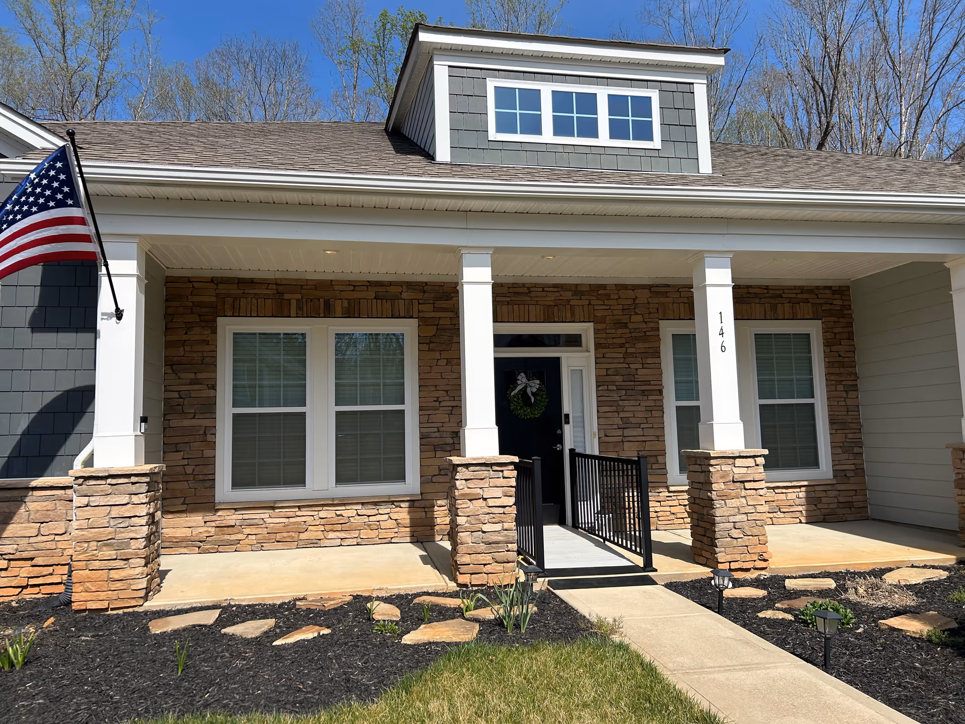 Front exterior view of a senior living facility unit with stone and siding facade, a covered porch with white columns, an American flag on the left, and a black door with a green wreath. The unit number 146 is displayed on one of the columns. There is a paved walkway leading to the entrance and landscaped garden beds with mulch and small plants.