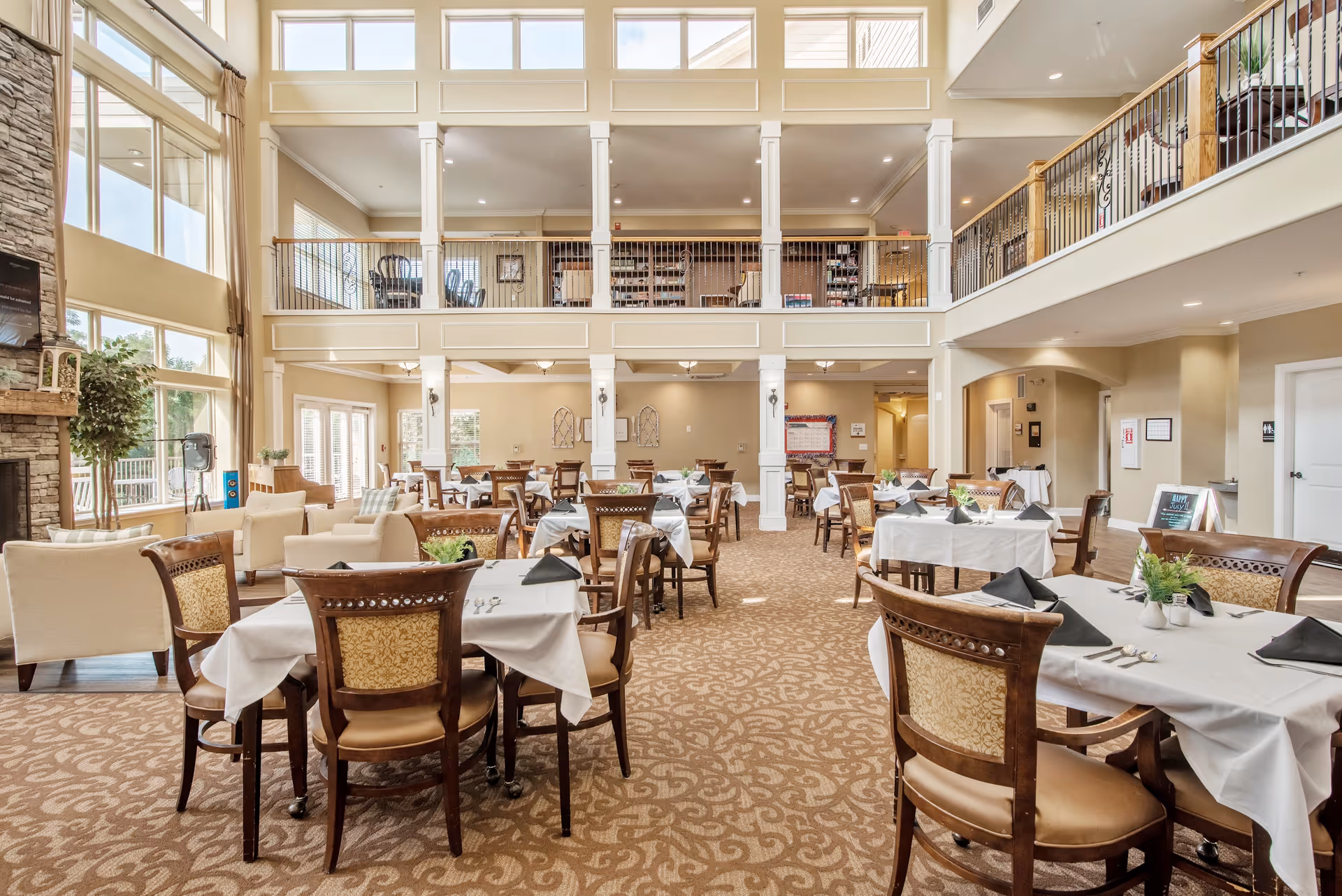 A spacious and well-lit dining room with multiple tables covered in white tablecloths and set with black napkins and silverware. The room features high ceilings with large windows allowing natural light to fill the space. There is a stone fireplace on the left side and a second-floor balcony overlooking the dining area. Comfortable chairs and some lounge seating are arranged around the tables.