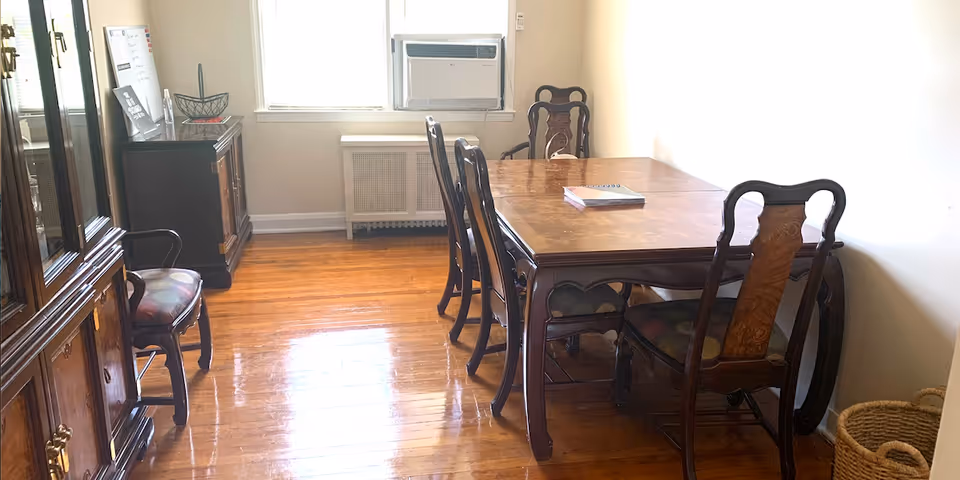 Wooden dining table with chairs in a bright, sparsely furnished dining room.