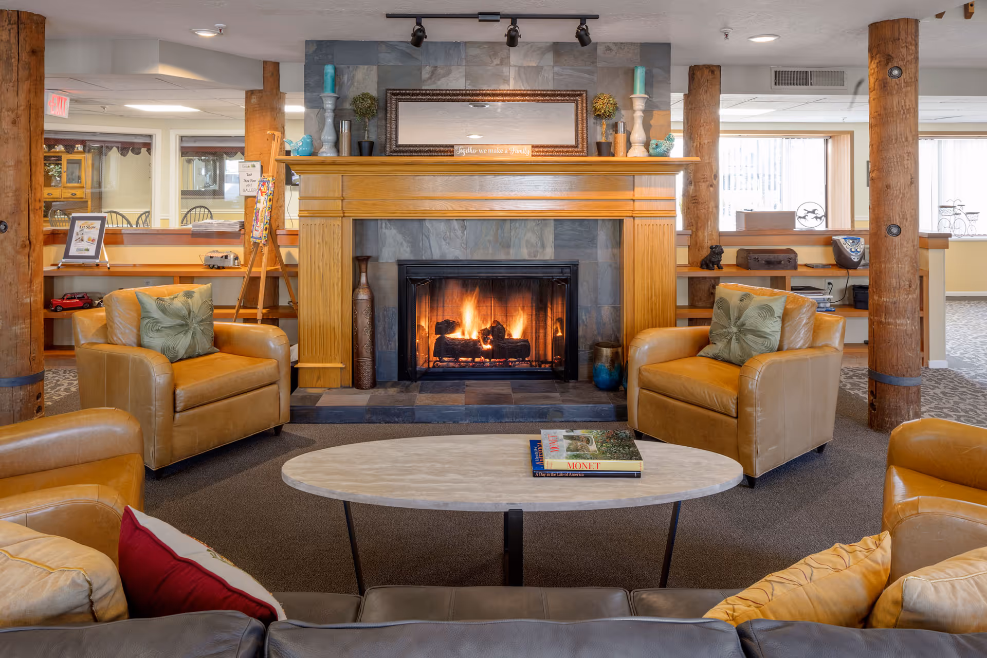 Cozy living room area with a lit fireplace surrounded by a wooden mantel and stone backdrop. The room features four leather armchairs arranged around an oval coffee table with books on it. The space has wooden support beams and shelves with decorative items, and large windows letting in natural light.