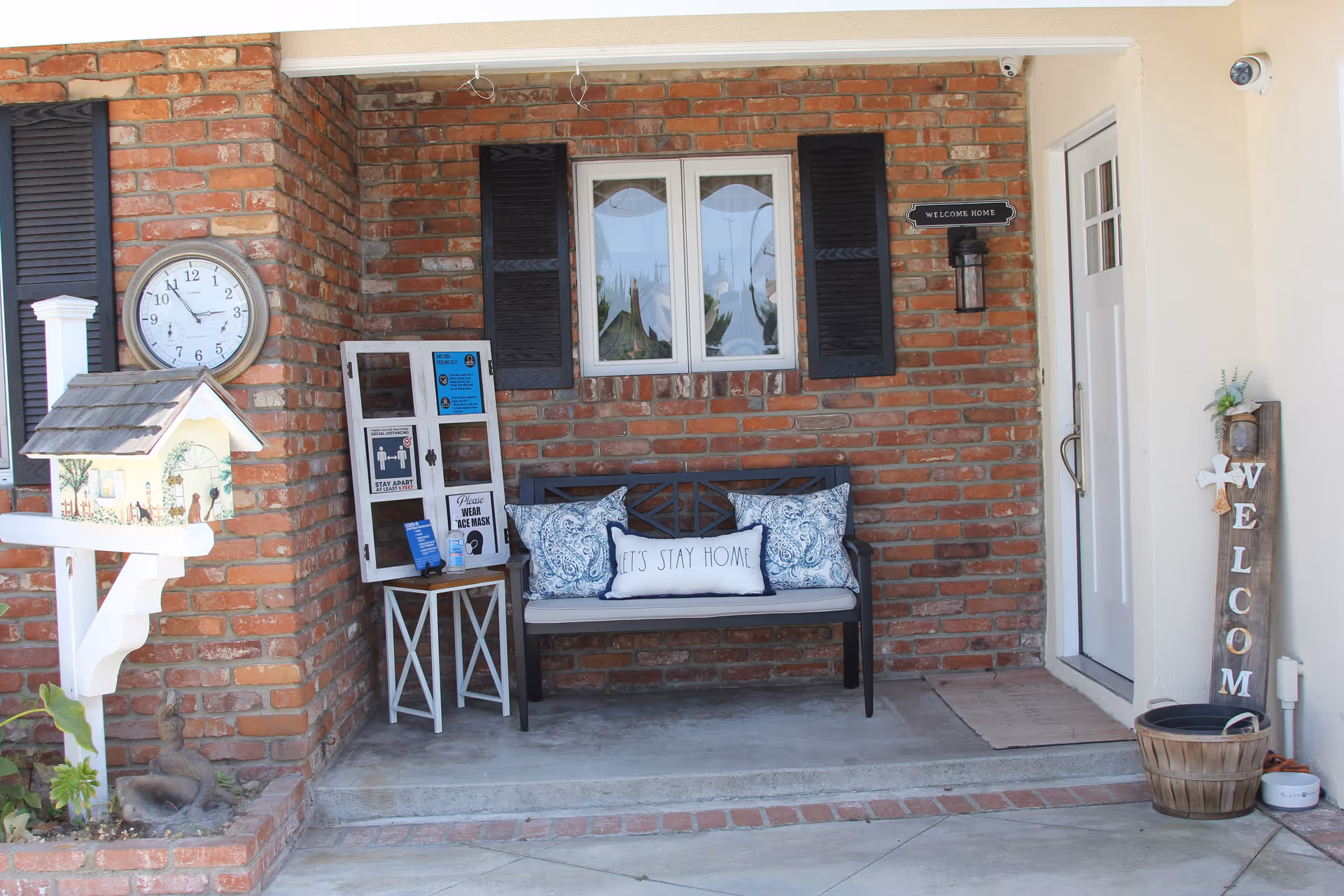 A cozy front porch area with a brick wall backdrop, featuring a bench with decorative pillows including one that says 'LET'S STAY HOME'. There is a white door to the right with a welcome sign beside it, a small table with informational signs, a wall clock, and a birdhouse mounted on a post. The area is clean and inviting.
