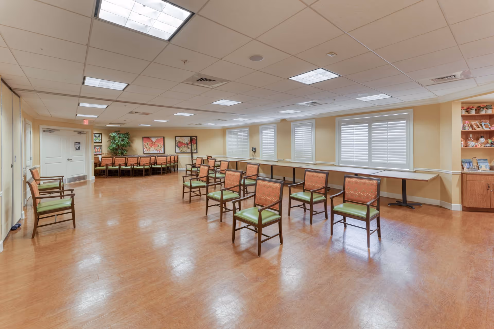 A spacious room with wooden flooring and beige walls, featuring multiple chairs with green seats and patterned backs arranged in rows and along the walls. The room has several windows with white shutters, fluorescent ceiling lights, framed butterfly artwork on the far wall, and a wooden cabinet with decorative items on the right side.